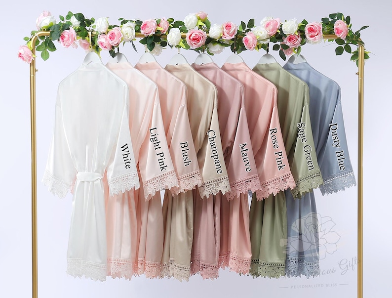 May include: A selection of satin robes in shades like white, pink, and blue, displayed on a gold rack. Each robe features lace detailing and a matching belt. The sleeves are labelled with colour names such as "White" and "Dusty Blue".
