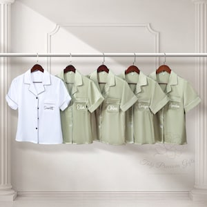 May include: Five personalized satin pajamas in sage green and one white satin pajama with black trim. The pajamas have short sleeves and a button-up collar. Each pajama has a name embroidered on the left chest pocket. The names are Scarlett, Ella, Chloe, Evelyn, and Amelia.