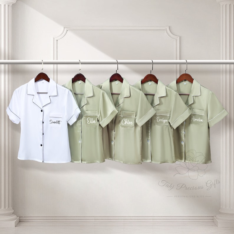 May include: Five personalized satin pajamas in sage green and one white satin pajama with black trim. The pajamas have short sleeves and a button-up collar. Each pajama has a name embroidered on the left chest pocket. The names are Scarlett, Ella, Chloe, Evelyn, and Amelia.
