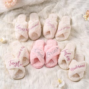 May include: A collection of fluffy, personalized slippers in cream and pink. The slippers are embroidered with names and titles like "Bride" and "Bridesmaid" in a cursive font. The slippers are arranged on a soft, white, textured surface.