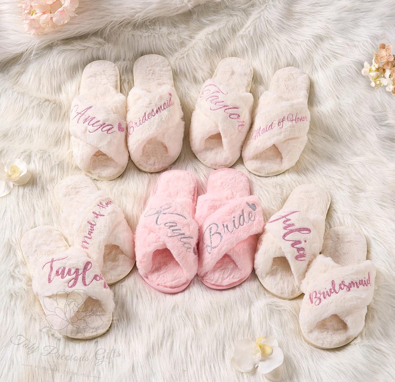May include: A collection of fluffy, personalized slippers in cream and pink. The slippers are embroidered with names and titles like "Bride" and "Bridesmaid" in a cursive font. The slippers are arranged on a soft, white, textured surface.