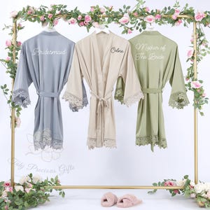 May include: Three silk robes in gray, beige, and olive green hang on a gold rack decorated with pink roses. Each robe has lace trim on the sleeves and hem. The gray robe says "Bridesmaid", the beige robe says "Celine", and the green robe says "Mother of The Bride".