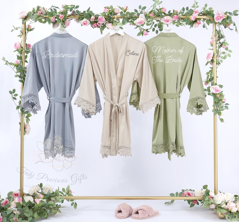 May include: Three silk robes in gray, beige, and olive green hang on a gold rack decorated with pink roses. Each robe has lace trim on the sleeves and hem. The gray robe says "Bridesmaid", the beige robe says "Celine", and the green robe says "Mother of The Bride".