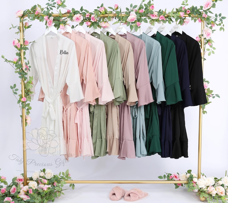 Puede incluir: Una colecci&oacute;n de batas de seda en varios colores, incluyendo blanco, rosa, verde y azul, colgadas en un estante dorado. Las batas tienen un dise&ntilde;o fluido con mangas anchas y cintura anudada. La palabra "Billie" est&aacute; bordada en una bata.