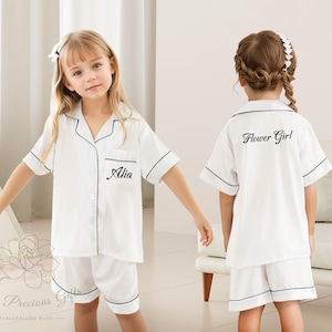 May include: Two girls wearing white pajamas with black trim. One pajama top has the text "Flower Girl" embroidered on the back. The other pajama top has the name "Alia" embroidered on the front pocket.