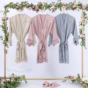 May include: Three satin robes in beige, mauve, and gray hang on a gold rack, each with lace trim and a matching belt. The robes are personalized with gold lettering. The rack is decorated with flowers and greenery.