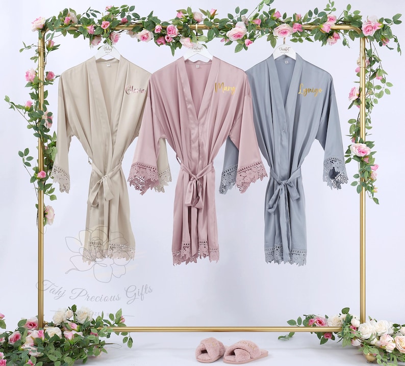 May include: Three satin robes in beige, mauve, and gray hang on a gold rack, each with lace trim and a matching belt. The robes are personalized with gold lettering. The rack is decorated with flowers and greenery.