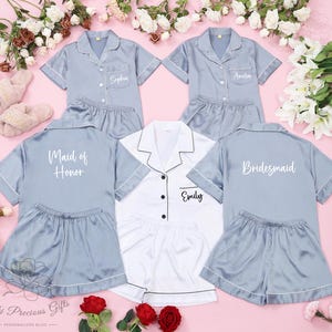 May include: Light blue satin pajama sets with white trim, personalized with names and titles like "Maid of Honor" and "Bridesmaid." A white pajama set is also included. The sets are displayed with flowers and a pair of fuzzy slippers.