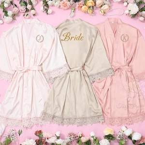 May include: Three satin bridal robes in blush pink and champagne colours, with lace trim and matching belts. One robe has "Bride" in gold script, and the others have initial monograms. Floral arrangement surrounds the robes.