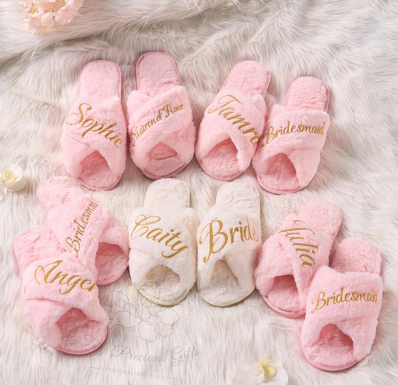 May include: A collection of pink and white fluffy cross-band slippers. Gold script lettering on the slippers includes names like "Sophie", "Tamra", and "Bride". The slippers are arranged on a white, textured surface.