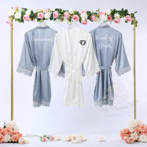 May include: Three satin robes on a gold rack, ideal for a bridal party. Two are a dusty blue colour, with the words "Bridesmaid" and "Maid of Honor" printed on the back. The third robe is white with a monogram. All robes have lace trim.