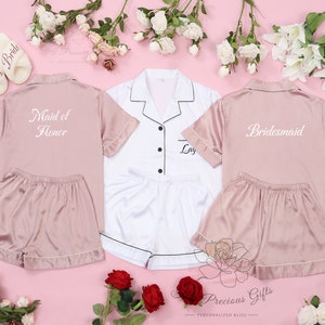 May include: Three satin pyjama sets in pink and white with short sleeves and shorts. The sets are personalised with the text "Maid of Honor", "Bride", and "Bridesmaid".