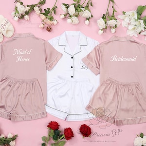 Puede incluir: Tres conjuntos de pijama de sat&eacute;n en rosa y blanco con mangas cortas y pantalones cortos. Los conjuntos est&aacute;n personalizados con el texto "Maid of Honor", "Bride" y "Bridesmaid".