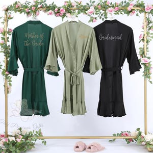 Puede incluir: Tres batas de seda en verde esmeralda, verde oliva y negro cuelgan de un estante dorado. La bata esmeralda dice "Mother of the Bride", la bata oliva "Allison" y la bata negra "Bridesmaid". Cada bata tiene cintur&oacute;n y mangas con volantes.