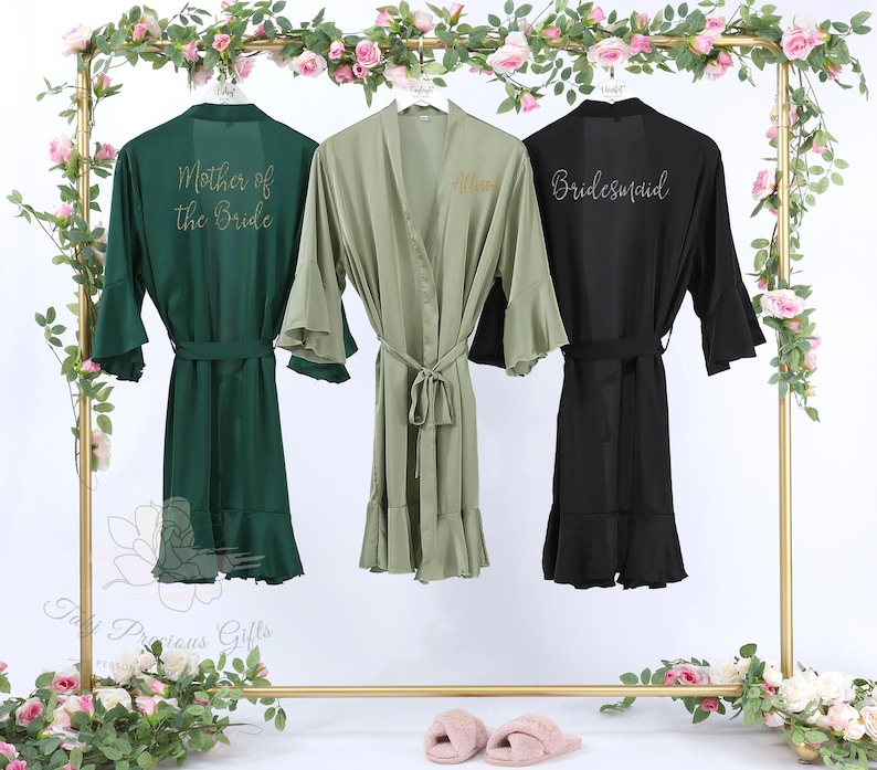 Puede incluir: Tres batas de seda en verde esmeralda, verde oliva y negro cuelgan de un estante dorado. La bata esmeralda dice "Mother of the Bride", la bata oliva "Allison" y la bata negra "Bridesmaid". Cada bata tiene cintur&oacute;n y mangas con volantes.