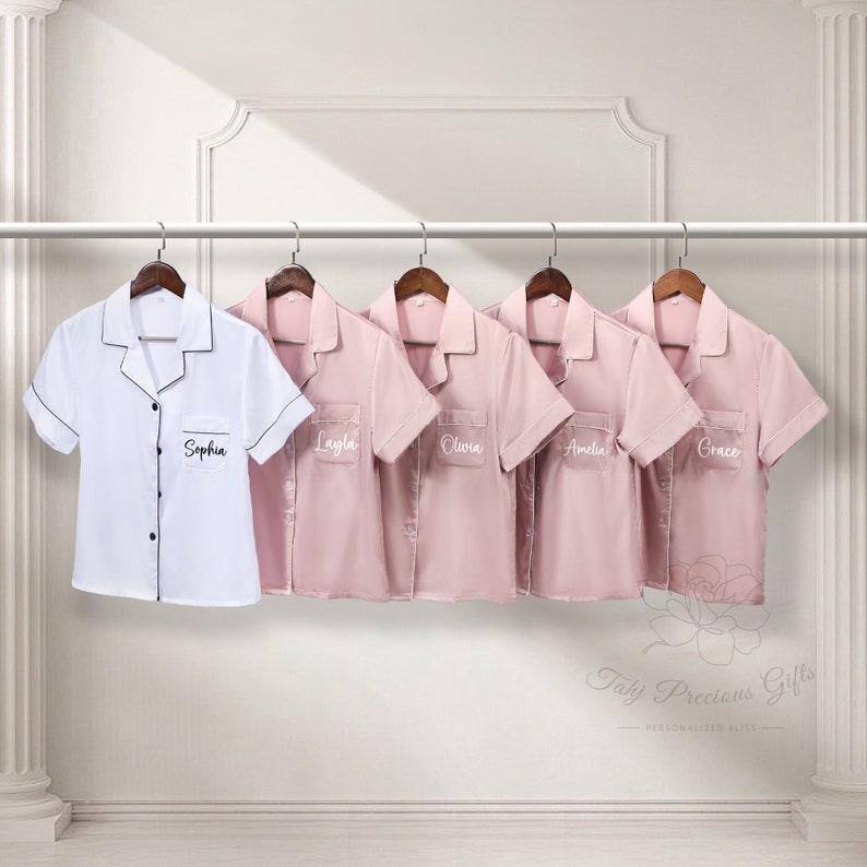 Puede incluir: Cinco camisas de pijama de manga corta colgadas en una barra. Una camisa blanca con ribete negro y el nombre "Sophia" en el bolsillo. Cuatro camisas rosas con los nombres "Layla", "Olivia", "Amelia" y "Grace" en los bolsillos.