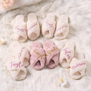 May include: A collection of plush, cross-band slippers in cream and pink. Each slipper is embroidered with names or titles like "Anya", "Bridesmaid", "Taylor", "Bride", and "Maid of Honor". The slippers rest on a fluffy white surface.