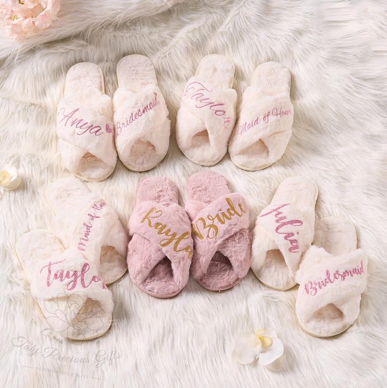 May include: A collection of plush, cross-band slippers in cream and pink. Each slipper is embroidered with names or titles like "Anya", "Bridesmaid", "Taylor", "Bride", and "Maid of Honor". The slippers rest on a fluffy white surface.