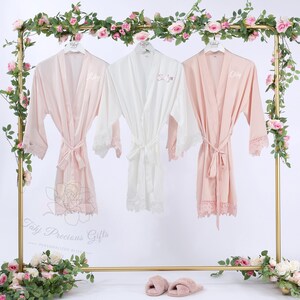 May include: Three silk robes in pink and white hang on a gold rack decorated with pink roses. The robes have lace trim and matching belts. Each robe is personalized with a name in cursive script. The rack is decorated with pink roses.