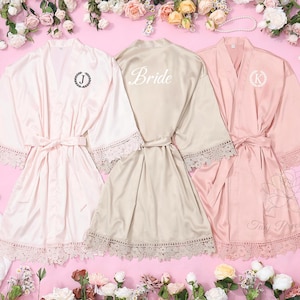 May include: Three satin robes in shades of pink and champagne, with lace detailing on the sleeves and hem. One robe features the word "Bride" on the back, and the others have initial monograms. Each robe is tied with a matching belt.