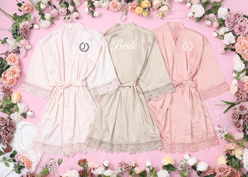 May include: Three satin robes in shades of pink and champagne, with lace detailing on the sleeves and hem. One robe features the word "Bride" on the back, and the others have initial monograms. Each robe is tied with a matching belt.