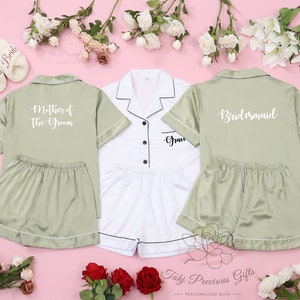 May include: Three satin pyjamas with short sleeves and shorts. One is white with black trim and the text "Groom" on the pocket. The other two are sage green with white trim. One says "Mother of the Groom" and the other says "Bridesmaid".