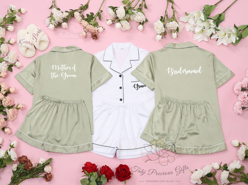 May include: Three satin pyjamas with short sleeves and shorts. One is white with black trim and the text "Groom" on the pocket. The other two are sage green with white trim. One says "Mother of the Groom" and the other says "Bridesmaid".