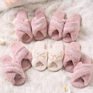 May include: A collection of pink and white fluffy cross-band slippers. Some slippers are embroidered with names like "Angel", "Bridesmaid", and "Maid of Honor". The slippers are arranged on a white, fluffy surface.