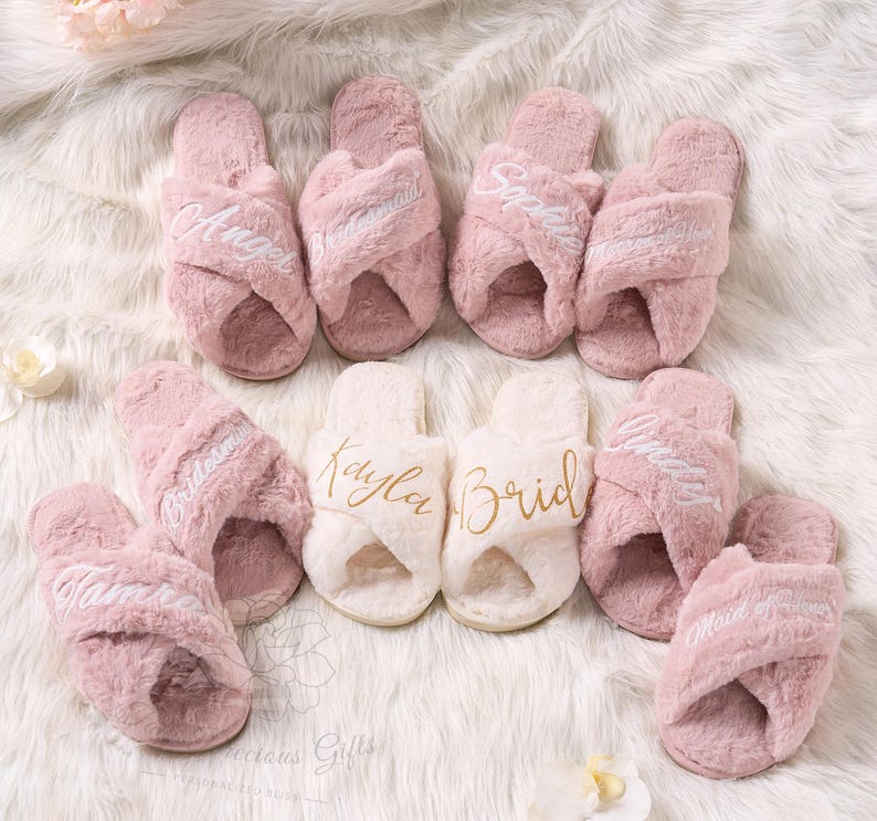May include: A collection of pink and white fluffy cross-band slippers. Some slippers are embroidered with names like "Angel", "Bridesmaid", and "Maid of Honor". The slippers are arranged on a white, fluffy surface.