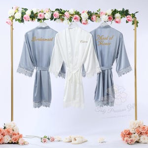 May include: Three satin robes hanging on a gold rack. Two are light blue, with "Bridesmaid" and "Maid of Honor" in gold lettering. The central robe is white, with the name "Catrin" in gold. Each robe has lace trim.