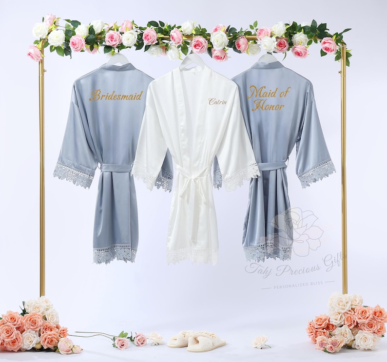 May include: Three satin robes hanging on a gold rack. Two are light blue, with "Bridesmaid" and "Maid of Honor" in gold lettering. The central robe is white, with the name "Catrin" in gold. Each robe has lace trim.
