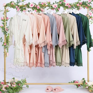 May include: A collection of silk robes in various colors, including ivory, peach, and navy, hang on a gold rack. Each robe features lace trim on the sleeves and a matching belt. The rack is decorated with pink roses and greenery, creating an elegant display.