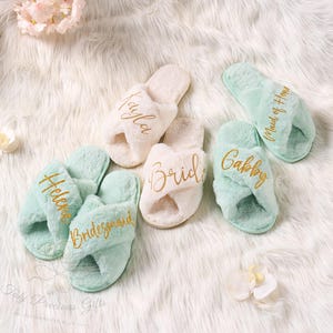 May include: A collection of fluffy, personalized slippers in white and mint green. Gold script embroidery includes names and titles like "Bride" and "Bridesmaid." The slippers are arranged on a soft, white, faux fur surface.