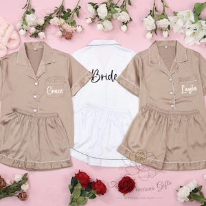 May include: Three satin pyjama sets with short sleeves and shorts. The sets are in a light brown colour with white piping. The sets are personalised with the names "Grace", "Bride", and "Layla" in white embroidery.