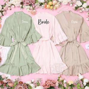 May include: Three satin robes in pastel shades: sage green, light pink, and beige. The green robe has the name "Sarina", the pink robe says "Bride", and the beige robe says "Gisela". Each robe has a matching belt and ruffled sleeves.