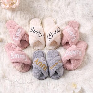 May include: A collection of fluffy, personalized slippers in various colors. The slippers are pink, cream, and gray, with embroidered names and titles like "Bride" and "Flower Girl". The slippers are arranged on a soft, white surface.
