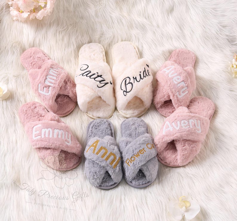 May include: A collection of fluffy, personalized slippers in various colors. The slippers are pink, cream, and gray, with embroidered names and titles like "Bride" and "Flower Girl". The slippers are arranged on a soft, white surface.