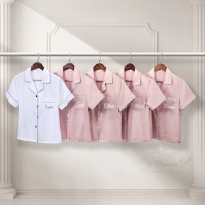 May include: Five personalised pajama shirts hanging on a rail. One white shirt with black trim and the name "Sophia" is on the left. The other four shirts are pink and have names embroidered on the pocket: "Layla", "Olivia", "Amelia", and "Emma".