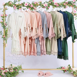 May include: A collection of satin robes in various colors, including white, pink, blue, and green, hang on a gold rack. Each robe has lace trim and a matching belt. The robes are labeled with color names like "White", "Light Pink", and "Forest Green".