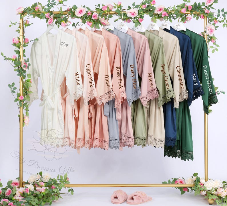 May include: A collection of satin robes in various colors, including white, pink, blue, and green, hang on a gold rack. Each robe has lace trim and a matching belt. The robes are labeled with color names like "White", "Light Pink", and "Forest Green".