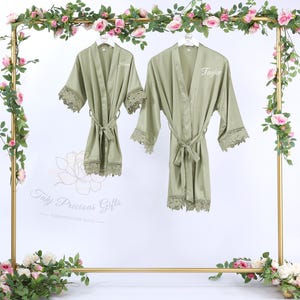May include: Two sage green robes with lace trim and matching belts hang on a gold rack decorated with pink roses and greenery. One robe is labeled "Allison" and the other "Taylor". The robes are part of the "Taly Precious Gifts" collection.