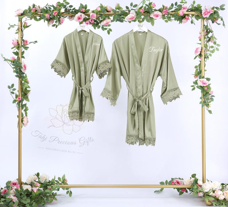 May include: Two sage green robes with lace trim and matching belts hang on a gold rack decorated with pink roses and greenery. One robe is labeled "Allison" and the other "Taylor". The robes are part of the "Taly Precious Gifts" collection.