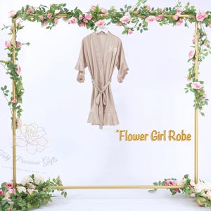 Puede incluir: Bata de sat&eacute;n beige con mangas y dobladillo con volantes, atada a la cintura. La bata cuelga de un marco dorado decorado con rosas rosas y vegetaci&oacute;n. El texto "Flower Girl Robe" es visible.