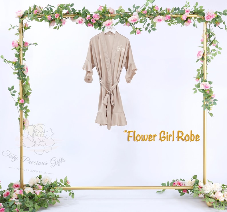 Puede incluir: Bata de sat&eacute;n beige con mangas y dobladillo con volantes, atada a la cintura. La bata cuelga de un marco dorado decorado con rosas rosas y vegetaci&oacute;n. El texto "Flower Girl Robe" es visible.