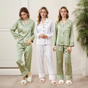 May include: Three women wearing satin pajama sets. One set is white with black trim, and the other two are a light green color. The pajamas have a button-down shirt and matching pants. The women are also wearing fuzzy slippers.