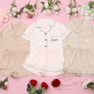 Puede incluir: Tres conjuntos de pijamas de sat&eacute;n personalizados en beige y rosa claro, con pantalones cortos a juego. Cada conjunto tiene un nombre bordado en la camisa. La imagen tambi&eacute;n incluye un par de zapatillas rosas y arreglos florales sobre un fondo rosa.