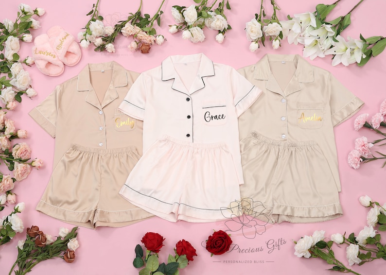 Puede incluir: Tres conjuntos de pijamas de sat&eacute;n personalizados en beige y rosa claro, con pantalones cortos a juego. Cada conjunto tiene un nombre bordado en la camisa. La imagen tambi&eacute;n incluye un par de zapatillas rosas y arreglos florales sobre un fondo rosa.