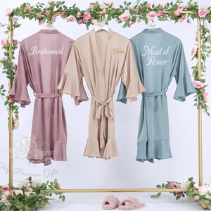 Puede incluir: Tres batas de sat&eacute;n en rosa empolvado, beige y verde azulado cuelgan de un estante dorado, adornadas con guirnaldas florales. La bata rosa empolvado dice "Bridesmaid", la bata beige tiene el nombre "Norma", y la bata verde azulado dice "Maid of Honor". Cada bata tiene un cintur&oacute;n a juego.