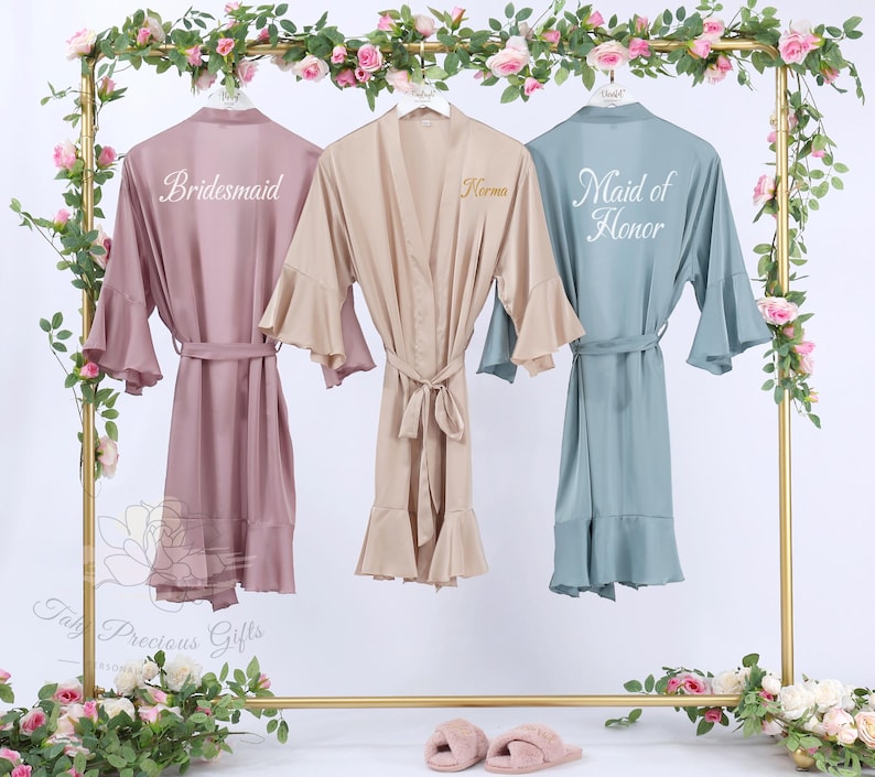 Puede incluir: Tres batas de sat&eacute;n en rosa empolvado, beige y verde azulado cuelgan de un estante dorado, adornadas con guirnaldas florales. La bata rosa empolvado dice "Bridesmaid", la bata beige tiene el nombre "Norma", y la bata verde azulado dice "Maid of Honor". Cada bata tiene un cintur&oacute;n a juego.