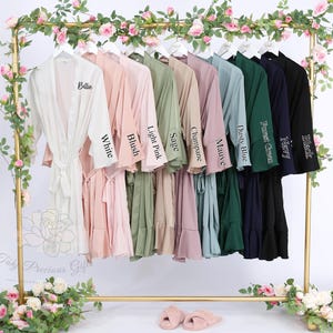 Puede incluir: Una exhibici&oacute;n de batas de varios colores colgadas en un estante dorado, incluyendo blanco, rubor, rosa claro, salvia, champ&aacute;n, malva, azul polvoriento, verde bosque, azul marino y negro. Cada bata tiene un cintur&oacute;n a juego y est&aacute; etiquetada con su nombre de color.
